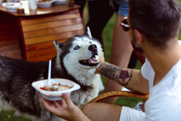 Young man and his dog at barbecue dinner on sunset time. Having meal outdoor in a forest glade or in backyard. Celebrating, relaxing. Summer lifestyle, food, resting, holidays, weekend, pet concept.