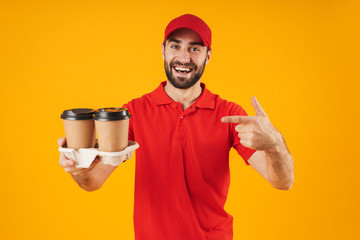 Wall Mural - Portrait of cheerful delivery man in red uniform smiling and holding takeaway coffee cups