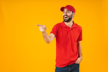 Wall Mural - Portrait of positive delivery man in red t-shirt and cap smiling and pointing finger at copyspace