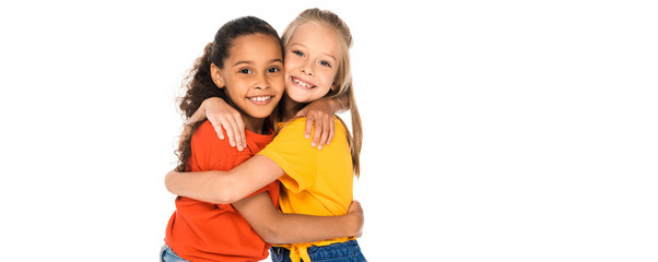 panoramic shot of two happy multicultural friends hugging while looking at camera isolated on white