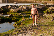 Seven-year-old Sports Smiling Boy Only In Swimming Trunks Is Barefoot With A Children's Bucket On The River