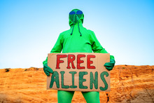A Man Dressed In A Green Alien Carnival Costume Suit At A Lone Rally With A Sign "free Aliens" On The Background Of A Desert Canyon