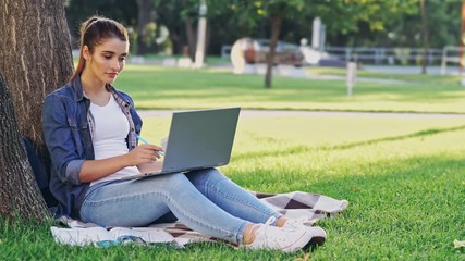 Poster - Smiling brunette woman using laptop computer and writing something while sitting near the tree on grass in park