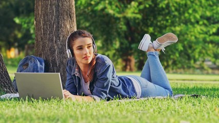 Poster - Pleased brunette woman using laptop computer and listening music while lying near the tree on grass in park