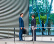 © Acronym - Confident business men talking in front of modern office building. Businessman and his colleague. Banking and financial market.