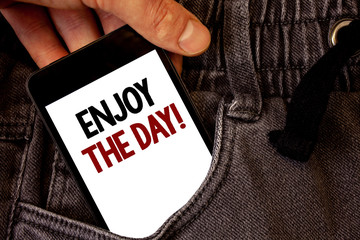 Text sign showing Enjoy The Day Motivational Call. Conceptual photo Enjoyment Happy Lifestyle Relaxing Time Text two words on white screen black Phone Hand holding grey jeans pocket