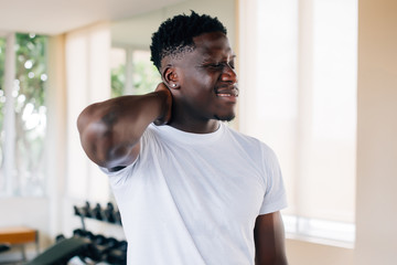 Fit African American man in white t-shirt holding sore neck and grimacing from pain on blurred gym background. Sportsman suffering from neck pain