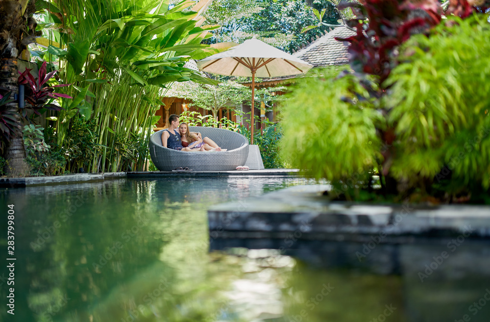 Candid shot of young caucasian couple lounging in modern chair near luxurious pool of tropical hotel and spa in Bali - obrazy, fototapety, plakaty 