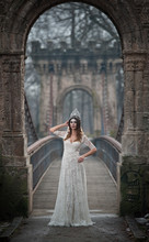 Lovely Young Lady Wearing Elegant White Dress Enjoying The Beams Of Celestial Light And Snowflakes Falling On Her Face. Pretty Brunette Girl In Long Wedding Dress Posing On A Bridge In Winter Scenery