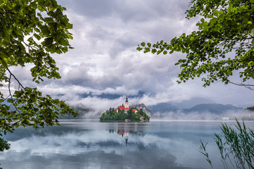 Wall Mural - Bled, Slovenia - Beautiful Lake Bled (Blejsko Jezero) with the Pilgrimage Church of the Assumption of Maria, Bled Castle and Julian Alps on a misty summer morning with summer foliage frame