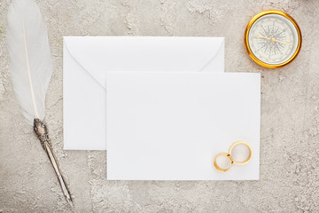 top view of wedding rings and quill pen on white blank card and golden compass on textured surface