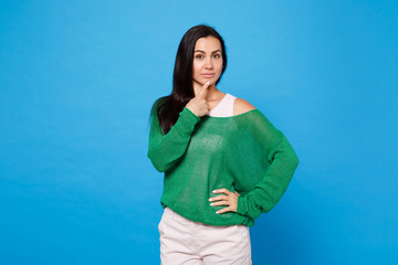 Portrait of attractive pensive young woman in green casual clothes looking camera, put finger prop up on chin isolated on blue wall background in studio. People lifestyle concept. Mock up copy space.