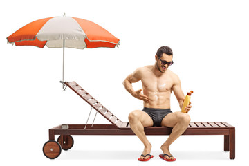 Wall Mural - Full length shot of a handsome fit man applying sun cream and sitting on a sunbed under umbrella isolated on white background