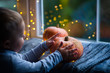 Toddler boy holding orange pumpkin on gray knitted plaid near window in evening surrounded with warm garland lights with golden bokeh.