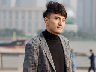 Portrait of a handsome Chinese young man in casual suit smiling and looking at camera confidently with Shanghai bund background, winter fashion, cool young man lifestyle.