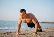 Concentrated sportsman with earphones doing push-ups at the beach in sunlight. Young beautiful man on the ocean coast.
