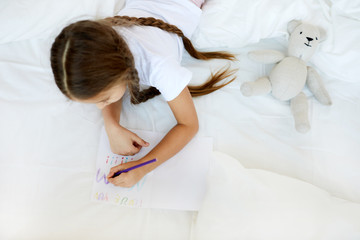 Wall Mural - Above view portrait of cute little girl drawing handmade card for Mothers day while laying in bed on white sheets, copy space