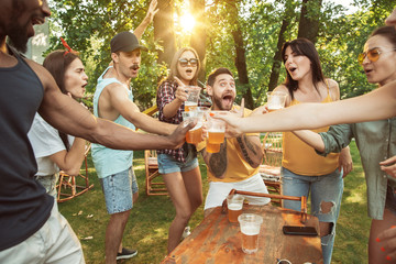 Group of happy friends having beer and barbecue party at sunny day. Resting together outdoor in a forest glade or backyard. Celebrating and relaxing, laughting. Summer lifestyle, friendship concept.