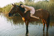 Woman lies astride a horse at river.