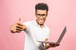 © denis_vermenko - Portrait of a happy young african man in casual holding screen laptop computer while showing thumbs up gesture and looking at camera isolated over pink background.