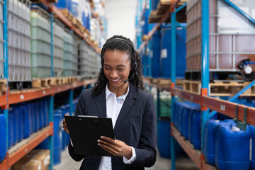 Female manager writing on clipboard while talking on headset in warehouse