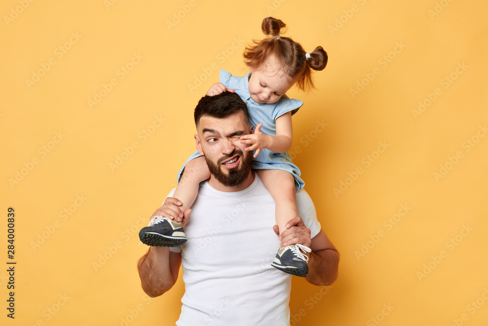 naughty behavior. parent cannot handle with kid with much energy. girl tiuching her dad's eyes while sittting on shoulders . close up photo. isolated yellow background. unacceptable kind of behaviour - obrazy, fototapety, plakaty 
