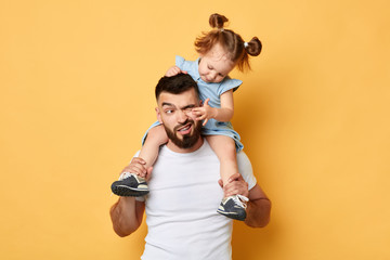 naughty behavior. parent cannot handle with kid with much energy. girl tiuching her dad's eyes while sittting on shoulders . close up photo. isolated yellow background. unacceptable kind of behaviour