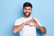 handsome cheerful guy is making a heart shape symbol with his fingers, expresses love and positive romantic feeling. Isolated on a blue background, positive man