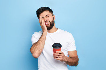 tired exhausted man covers face with palm, holds disposable paper cup of coffee. man doesn't want to