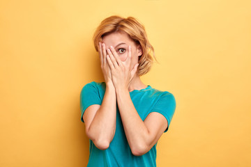 charming pleasant woman hiding her face , being afraid of something, looking through fingers to the camera, isolated yellow background, studio shot