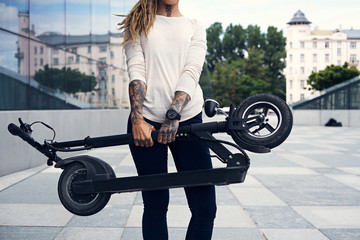 Wall Mural - Young tattooed woman with dreadlocks is posing for photographer while holding folded electro scooter.