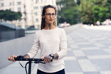 Wall Mural - Beautiful trendy girl with dreadlocks is driving electro scooter on the street in cloudy summer day.