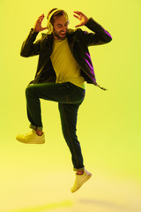 Wall Mural - Emotional handsome young stylish man jumping dancing isolated over light green background wall with led neon lights dressed in leather jacket listening music with headphones.