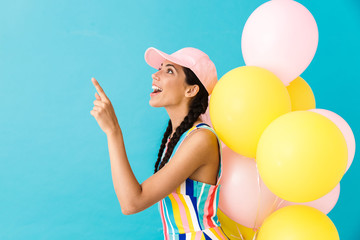 Wall Mural - Image of young amazed woman wearing cap pointing finger at copyspace while holding air balloons