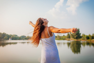 Young woman feeling free and happy raising arms and spinning around by summer river at sunset