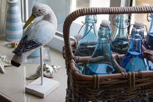 Close-up Of Several Glass Water Bottles In A Wicker Basket And A Decorative Wooden Bird On The Windowsill, Selective Focus