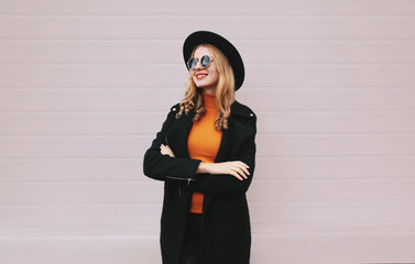 Wall Mural - Stylish smiling woman crossed her arms in black coat, round hat standing on gray wall background