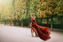 Mysterious Blond Woman In A Red Evening Dress, Walks In The Autumn Park Barefoot. The Wind Blows A Long Train And Hair. Queen In Her Garden. Rear View, Shooting From The Back Without A Face