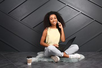 Wall Mural - Portrait of beautiful African-American woman with laptop talking by mobile phone near dark wall