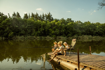 Cute little girls and their granddad are on fishing at the lake or river. Resting on pier near by water and forest in sunset time of summer day. Concept of family, recreation, childhood, nature.