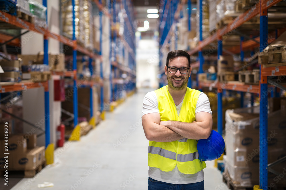 Portrait of middle aged warehouse worker standing in large warehouse distribution center with arms crossed. In background shelves with goods. - obrazy, fototapety, plakaty 