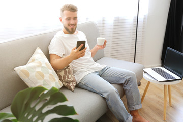 Bearded guy wearing blank white t-shirt & denim pants sitting alone at home on grey textile couch. Young man w/ facial hair in domestic situations. Interior background, copy space, close up, monstera.