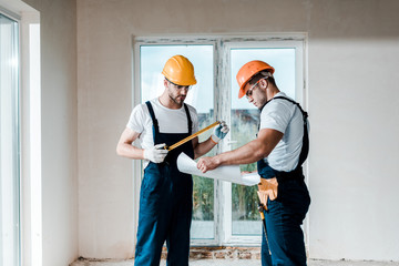 Wall Mural - handsome builder looking at blueprint near coworker holding measuring tape