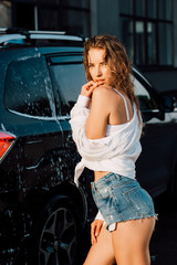 sexy young woman touching face and looking at camera near wet car