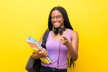 African American teenager student girl with long braided hair over isolated yellow wall points finger at you