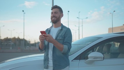 Handsome joyful man using smartphone leaning on car outdoor. Happy smiling male driver messaging on mobile phone looking around staying in modern city. Summertime.