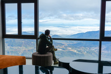 Hotel Room With Mountain Landscape View From Window, Interiors With Chairs And Table, And A Man Sits Seeing The Scene Through Window