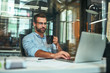 © Friends Stock - Morning coffee. Portrait of young and successful bearded man in eyeglasses holding cup of coffee and working with laptop while sitting at his working place