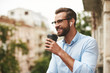 © Friends Stock - Positive news. Young and handsome bearded man in eyeglasses and headphones holding cup of coffee and talking with friend while standing at the office balcony