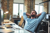 Fototapeta  - Enjoying break. Satisfied young bearded businessman leaning back with hands behind head and relaxing while sitting in the modern office